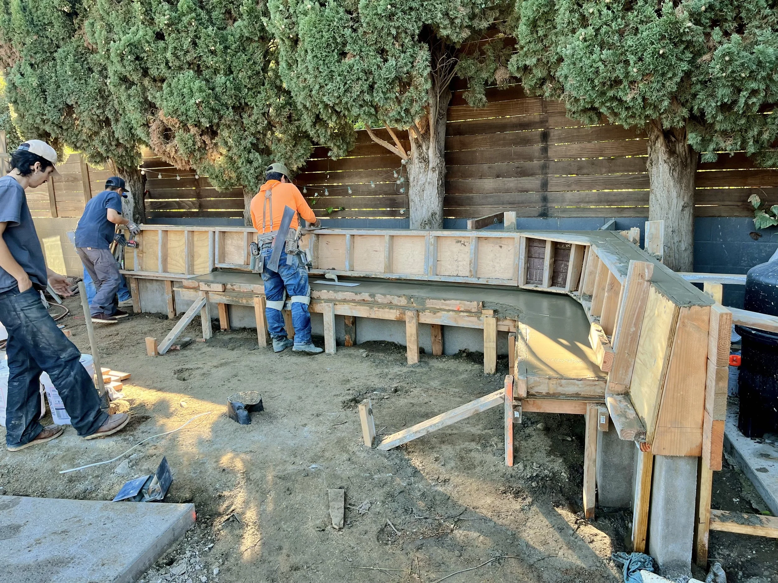Construction workers are building a wooden framed structure in a backyard with large trees and a wooden fence in the background.