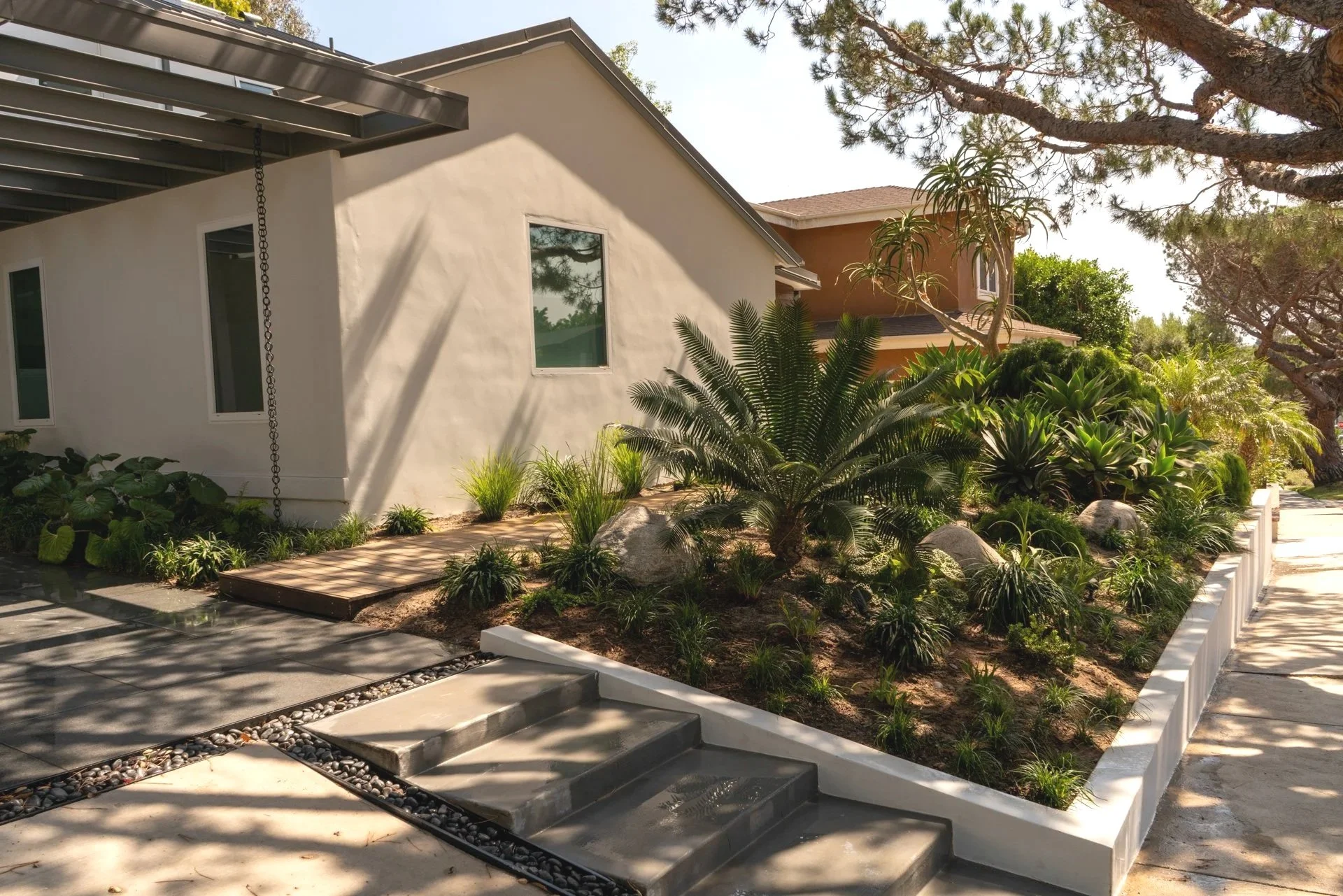 A modern house with white walls, large windows, and a sloped roof, surrounded by lush green plants and trees. A driveway with minimalist design features leads up to the house, and there is a staircase with a pebble-lined border in the front yard.