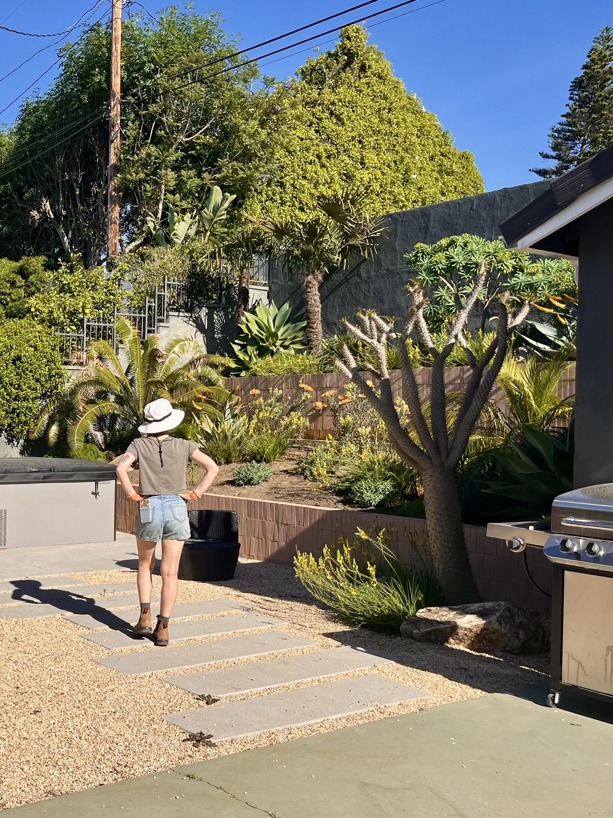 A woman with a wide-brimmed white hat, gray shirt, and denim shorts stands in a backyard garden, facing away. The garden features various plants, small trees, and a barbecue grill on the right. There are concrete stepping stones in the gravel pathway, a black basket, and a black chair.