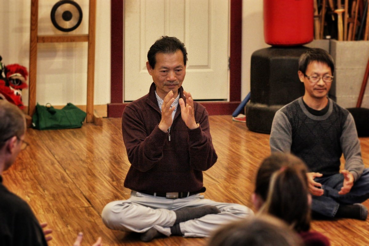 A group of people sitting cross-legged on a wooden floor, listening to a man in a maroon sweater who is sitting in a similar position with his hands raised, in what appears to be a meditation or discussion circle in a room.