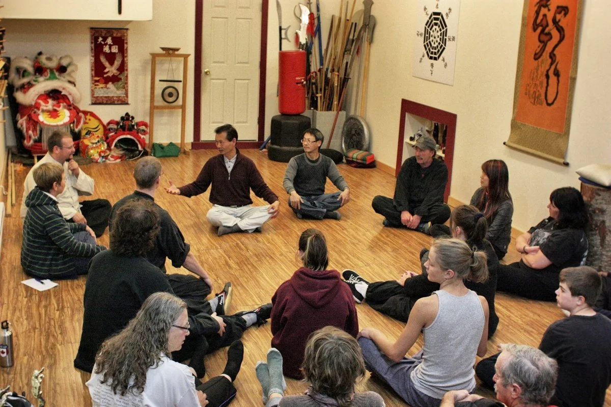 People sitting in a circle on a hardwood floor in a room decorated with Chinese art and artifacts, engaging in a group discussion or meditation session.
