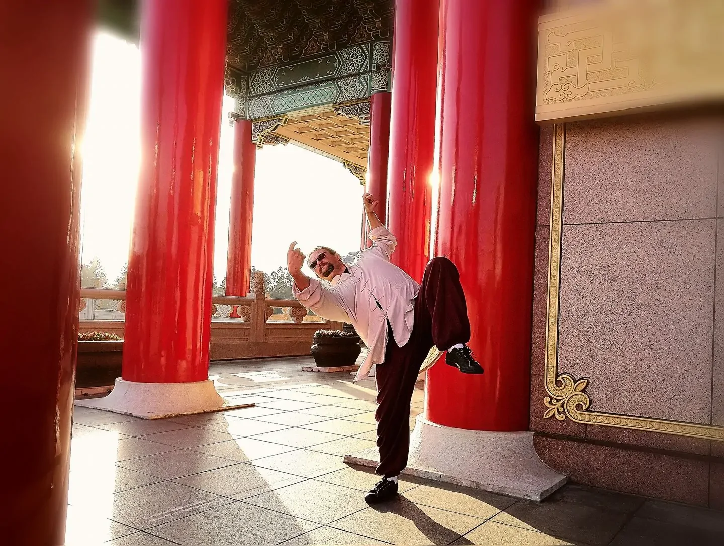 A man wearing sunglasses, a white jacket, and black pants is striking a playful pose near large red pillars at a temple or similar structure, with sunlight shining behind him.