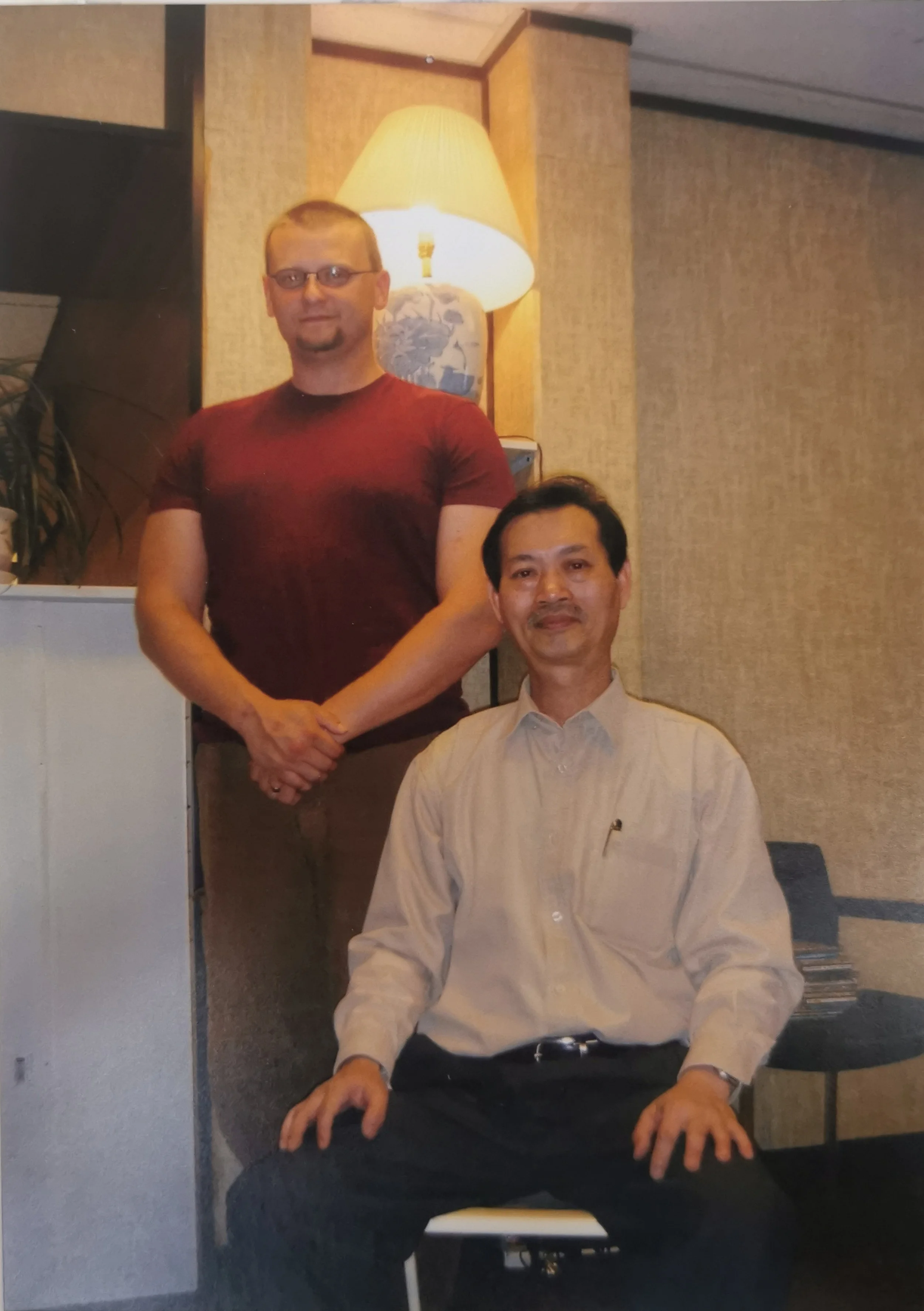 Two men, one sitting and one standing, in an indoor setting with beige walls and a lamp in the background.