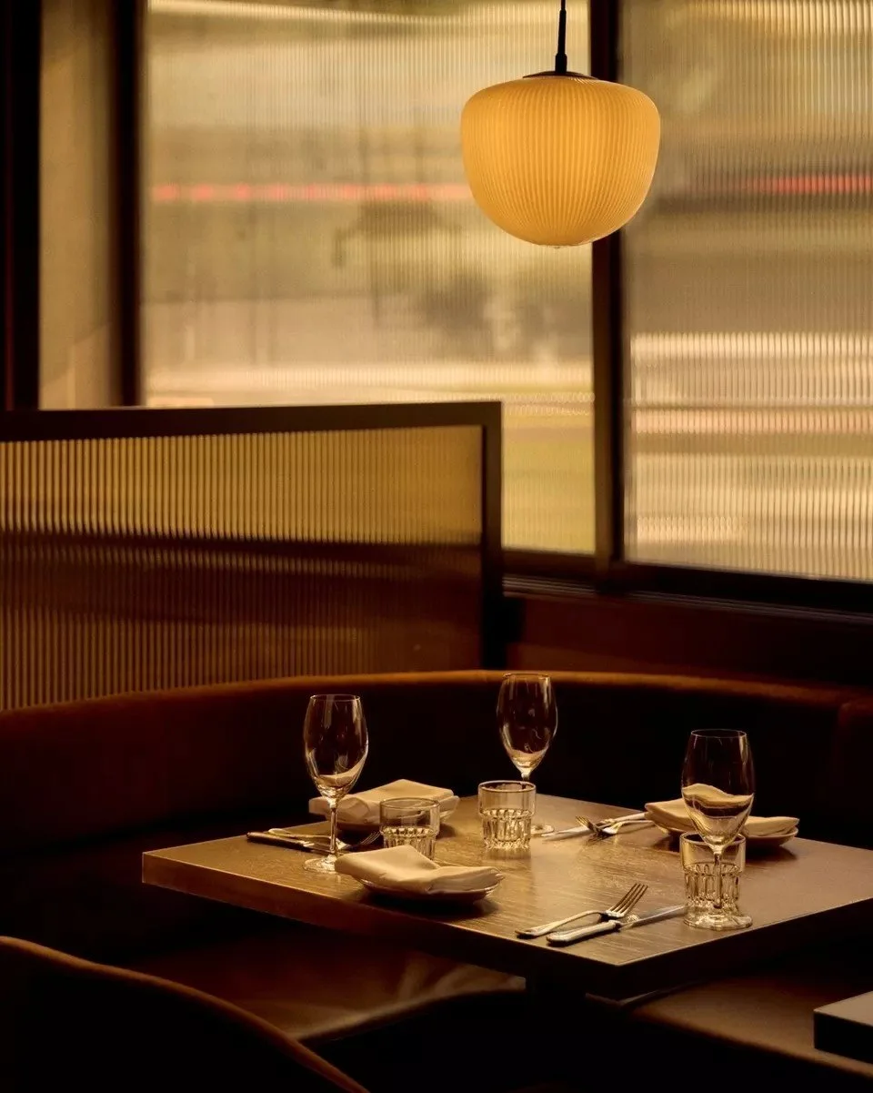 A restaurant table set with four wine glasses, four water glasses, four plates, and cutlery, illuminated by warm lighting with a hanging lamp, in a softly lit dining area with large windows.