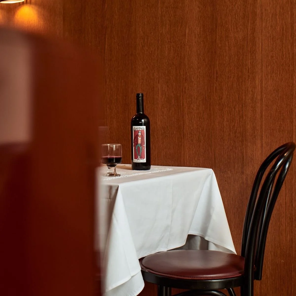 A table with a white tablecloth in a restaurant, holding a red wine bottle with a colorful label, a glass of red wine, and a wooden chair against a wood-paneled wall.
