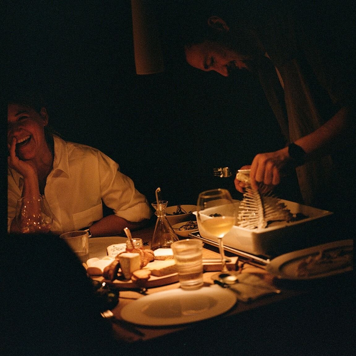 A woman laughing and a man working in a dimly lit, cozy dining setting with food and drinks on the table.