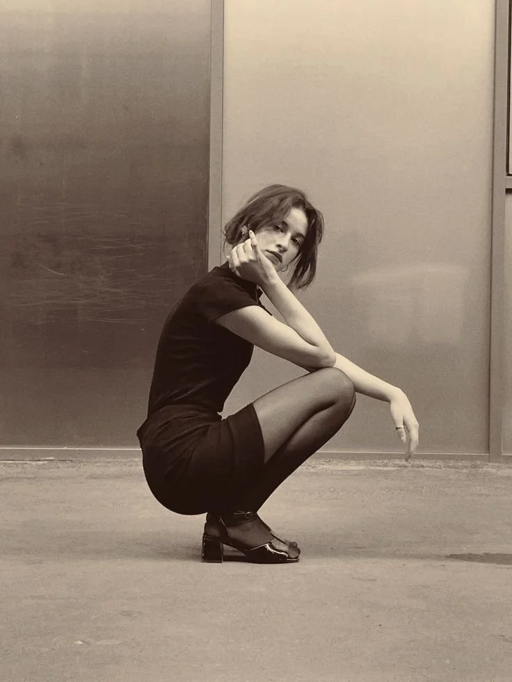 A woman with short hair squats on a floor in an indoor setting, wearing dark clothing and high-heeled shoes, looking at the camera.