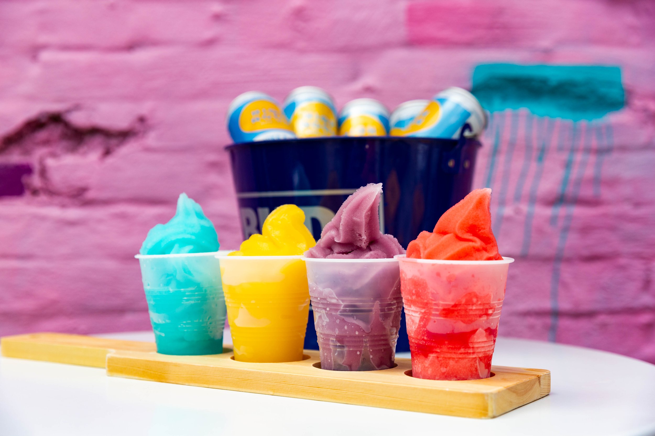 Six colorful cups of ice cream in a wooden ice cream holder, with a pink background and a blue container of small egg-shaped candies in the background.