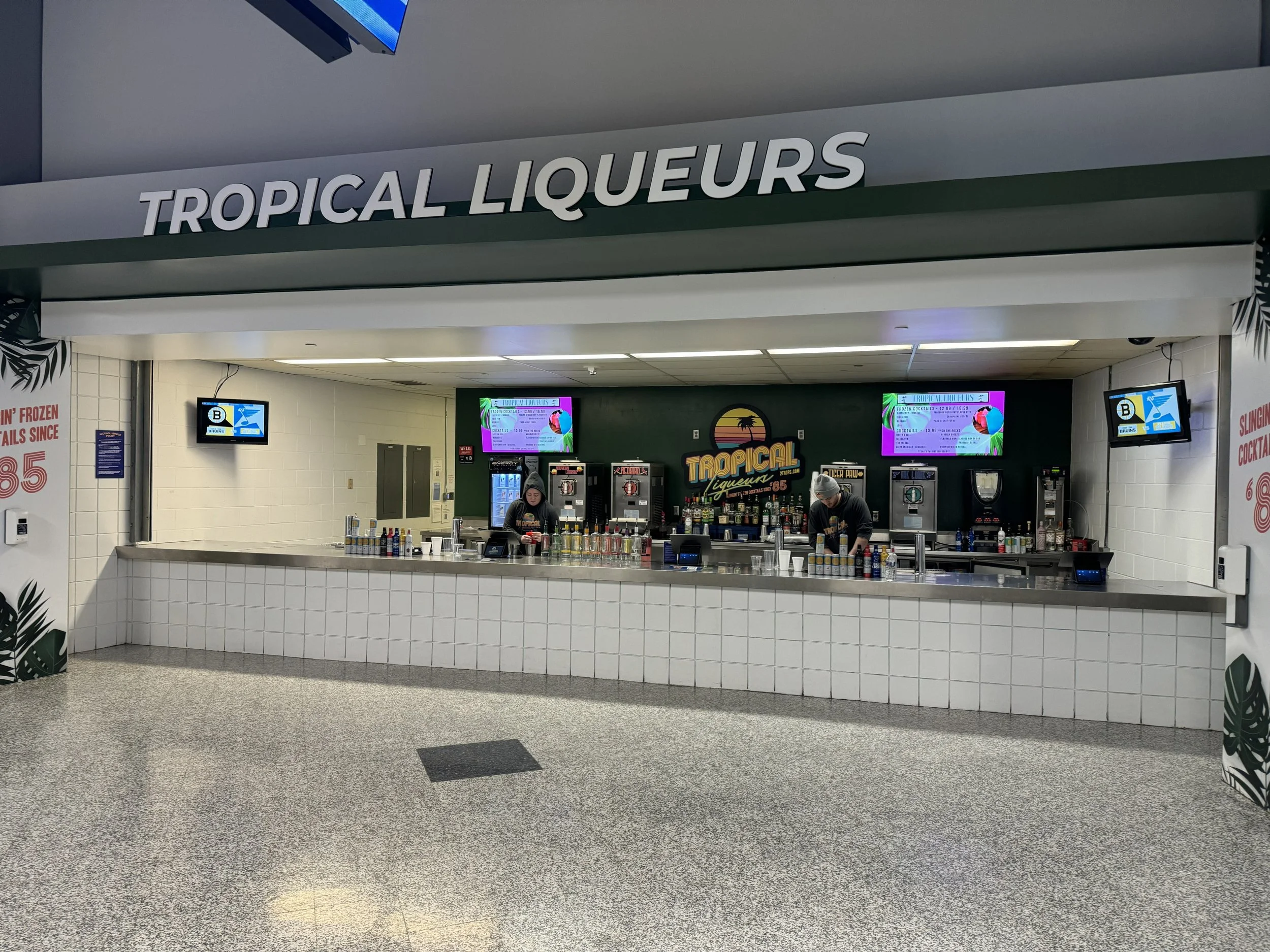 A food court or concession stand area with a sign that reads 'Tropical Liqueurs'. The stand has two digital menu boards displaying drinks, and a few staff behind the counter preparing beverages. The surroundings show a tiled front counter and a few people working.