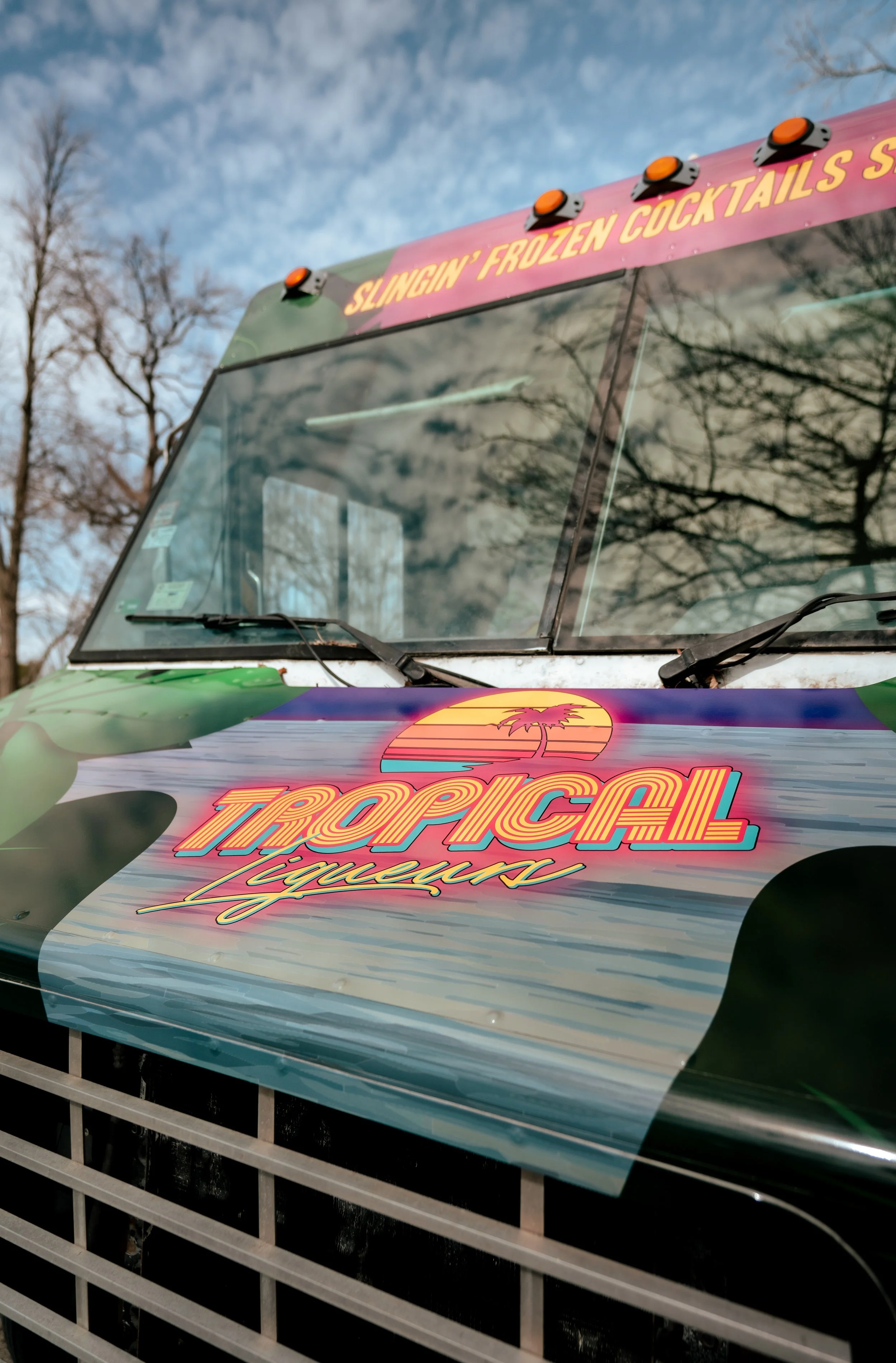 Close-up of a food truck with vibrant tropical-themed signage, including a pink banner that reads 'Slingin' Frozen Cocktails' and a colorful logo featuring a sunset and palm tree with the words 'Tropical Liquors,' behind a metal grill.