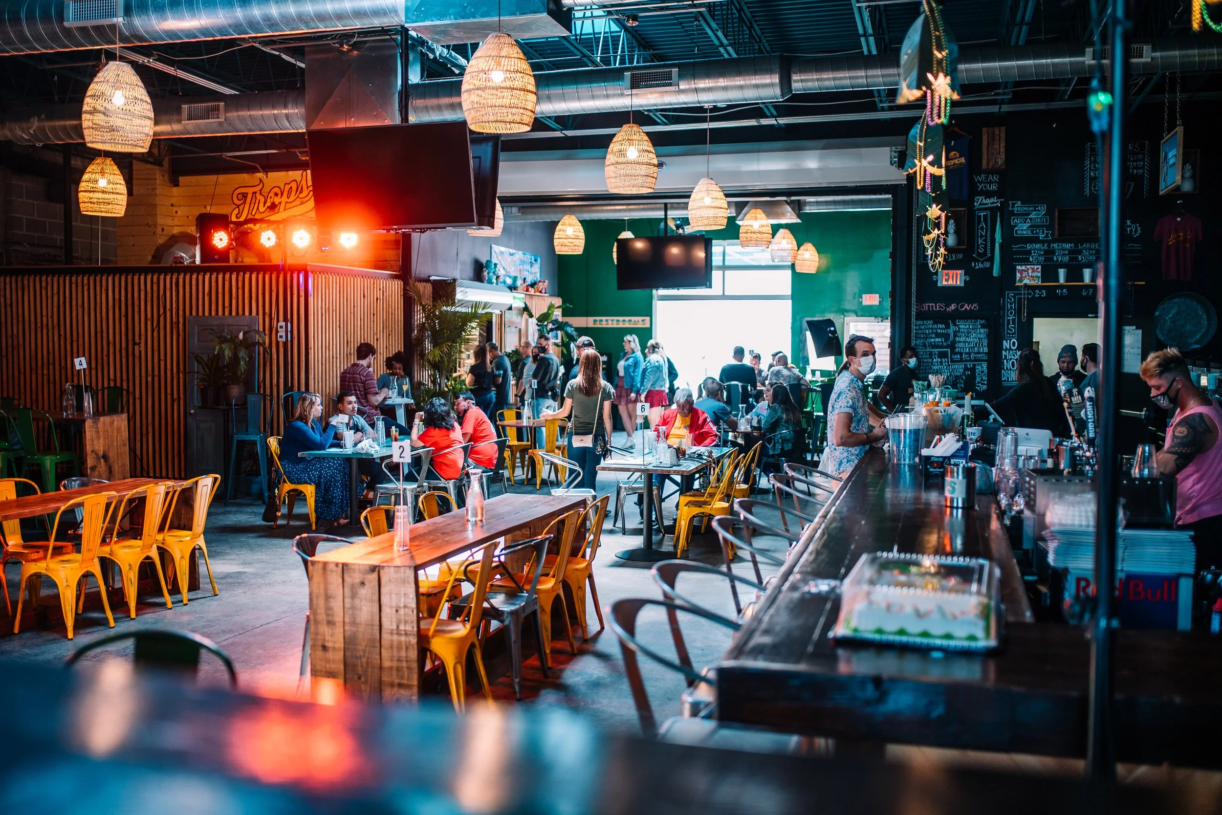Indoor bar and restaurant filled with people, with hanging lantern-style lights, a bar counter with bartenders, and tables with patrons, some wearing face masks, in a lively atmosphere.
