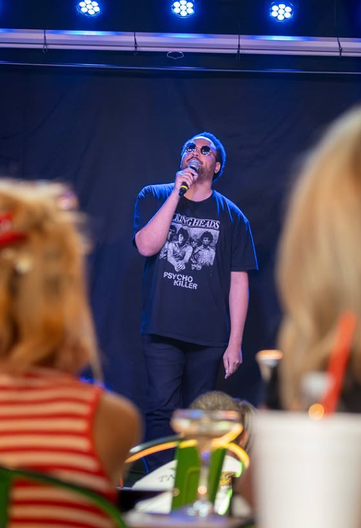 A man wearing sunglasses and a black T-shirt with band photos and text, holding a microphone, singing on stage with blue overhead lights.