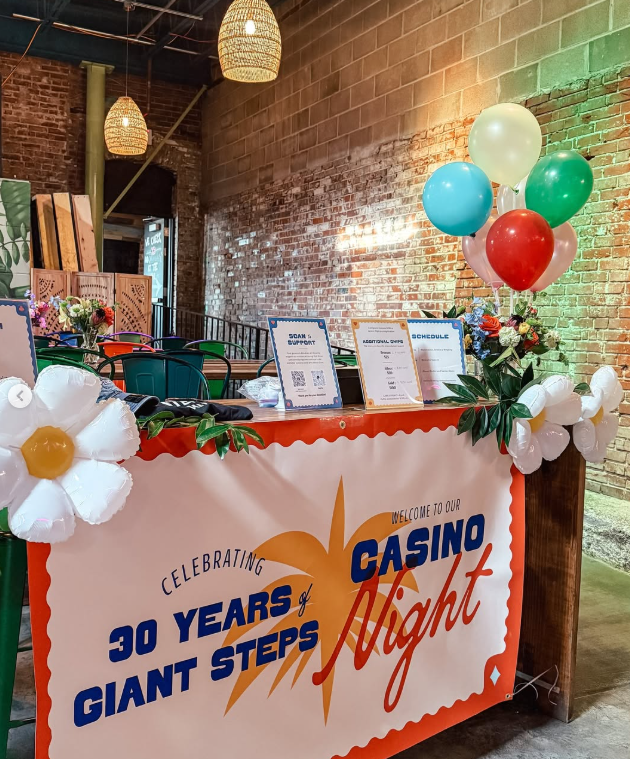 Event table decorated with balloons and flowers for a casino night celebrating 30 years of Giant Steps, with informational signs and a welcoming banner.