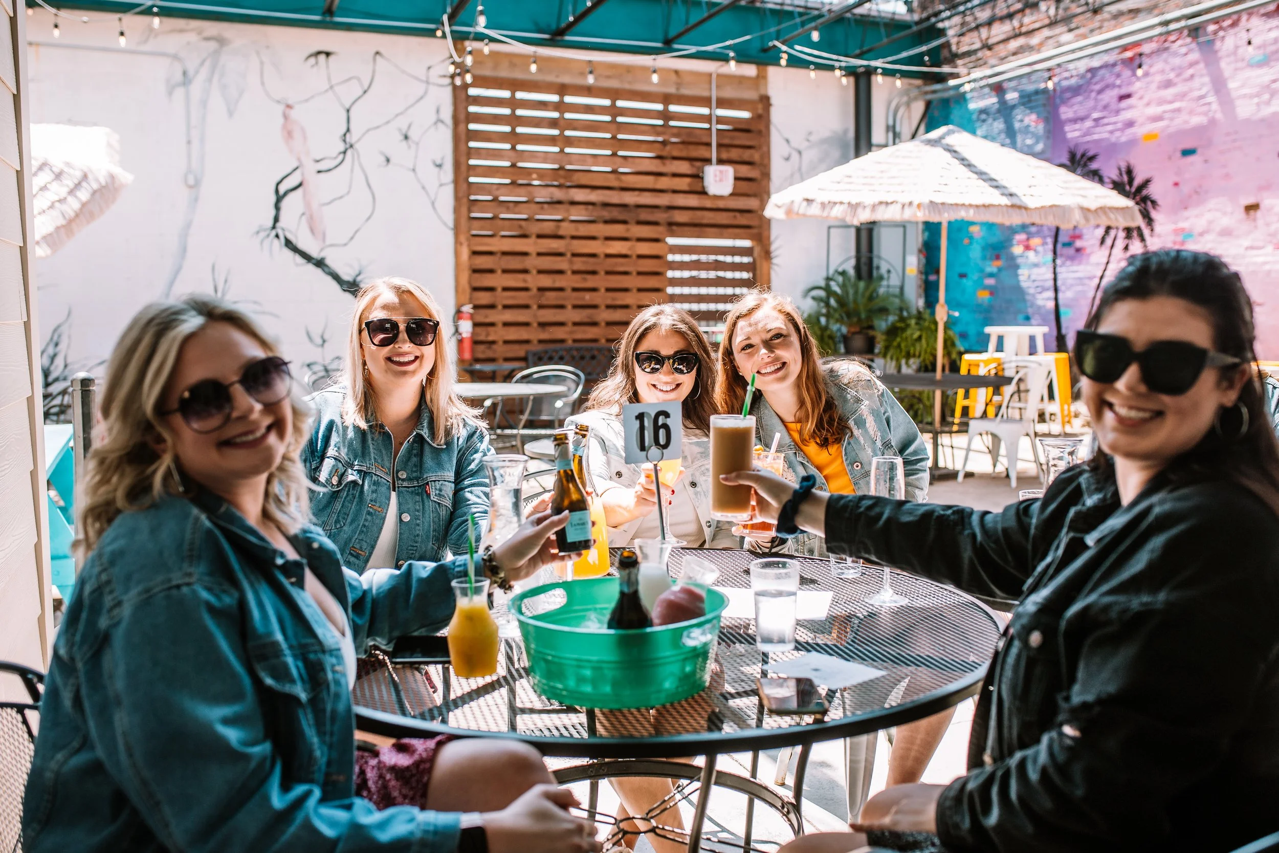 A group of smiling women wearing sunglasses sitting at an outdoor table in a colorful patio, enjoying drinks and celebrating together.