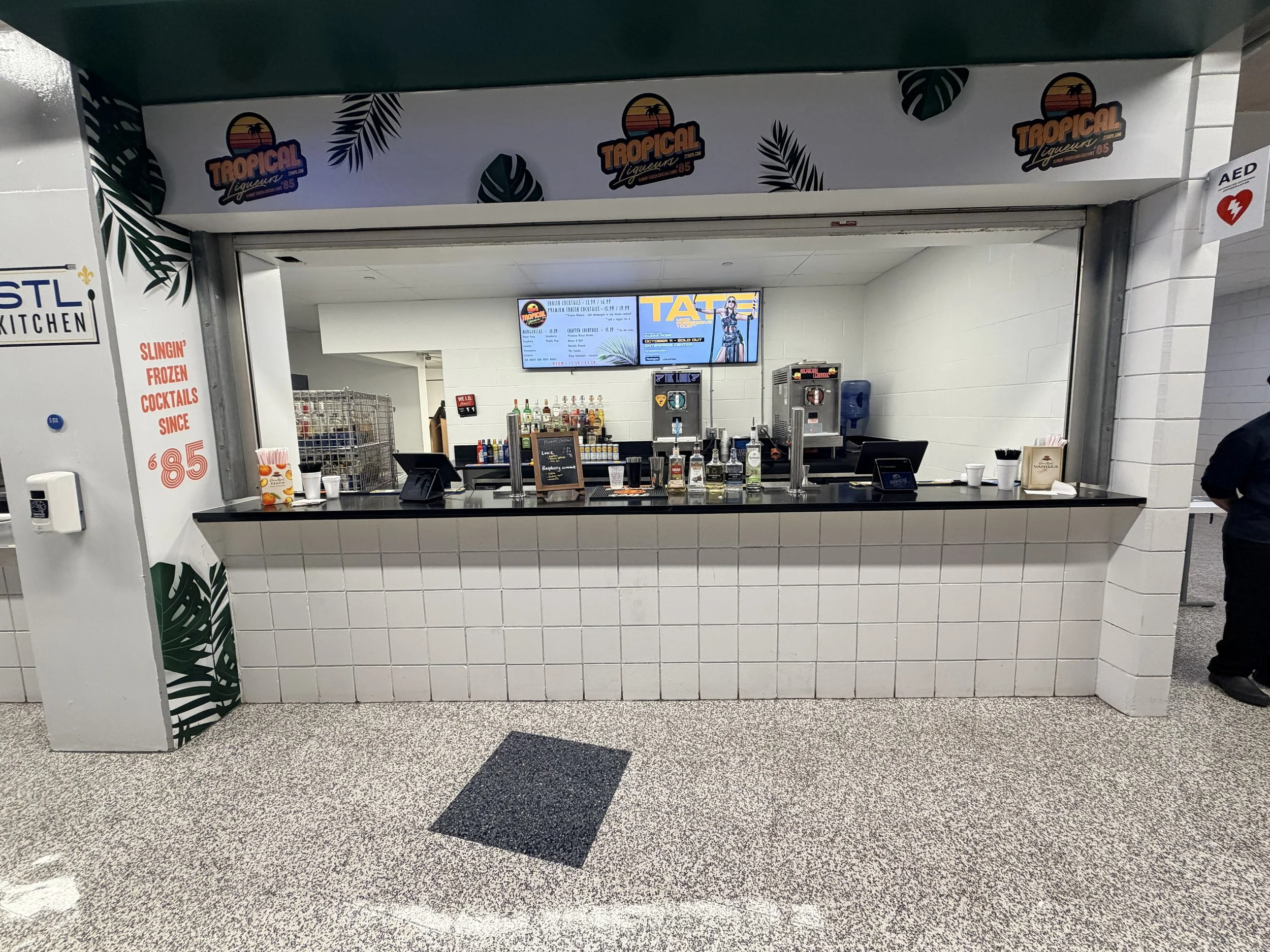 Bar kiosk with liquor bottles and a digital menu, located in an airport or similar public space, with a tropical theme including palm tree graphics, and a sign advertising frozen cocktails since $6.85.