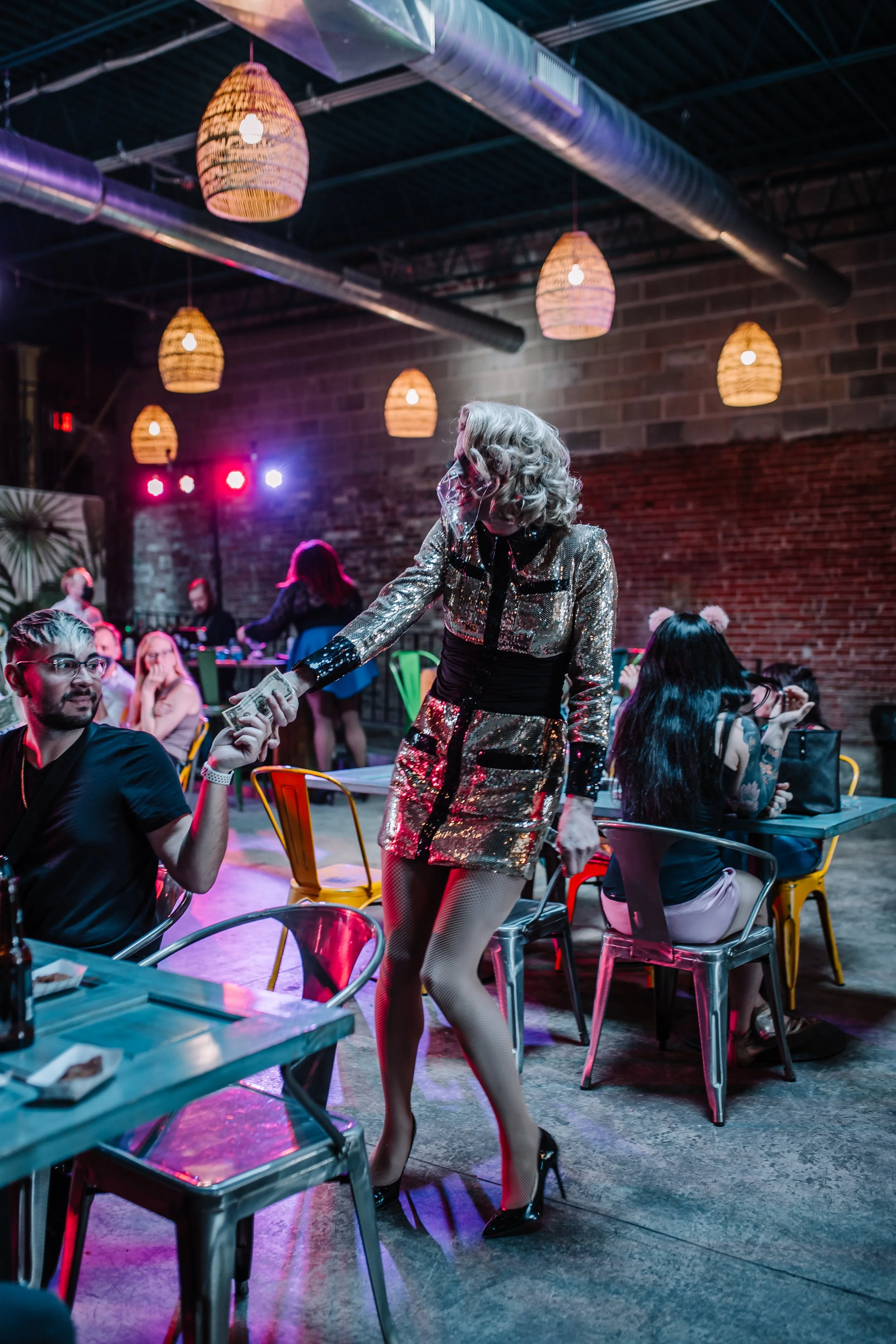 A woman dressed in a shiny, sequined outfit with fishnet tights and high heels is shaking hands and exchanging money with a seated man in a crowded bar or nightclub with colorful lighting and hanging lamps.