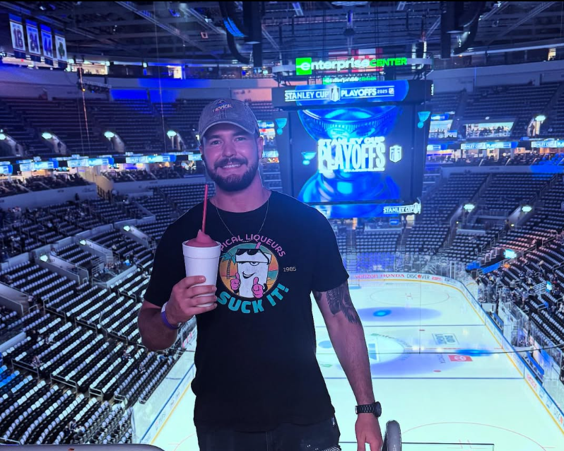 A man with a beard and baseball cap smiling at the camera, holding a drink in a cup with a straw, inside an empty hockey arena with a large scoreboard reading 'Playoffs' and 'Stanley Cup' in the background.