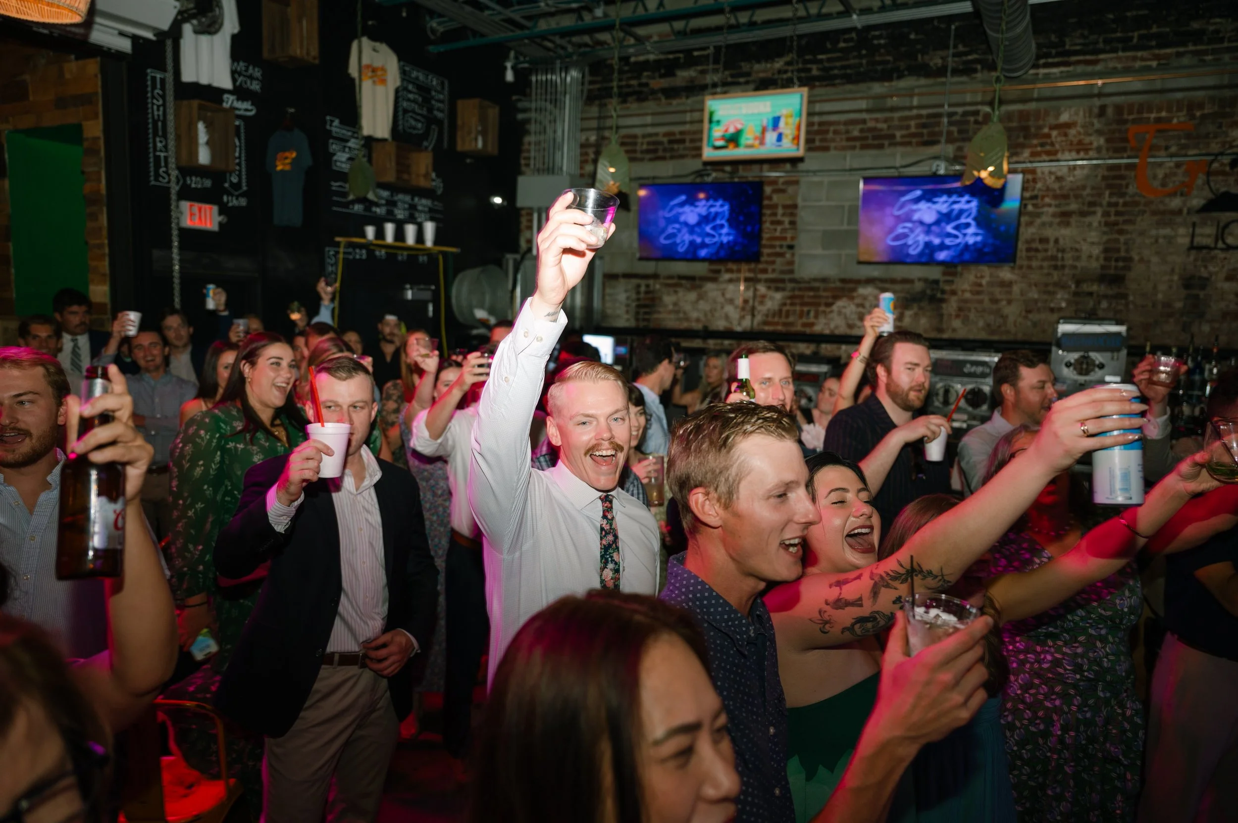 Crowd of enthusiastic people raising glasses and drinks at a lively bar or nightclub celebration with brick walls and multiple TVs displaying neon-colored graphics.