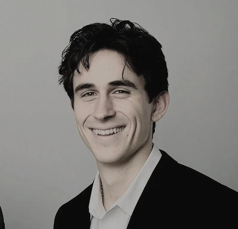 Portrait of a young man with dark hair, smiling, wearing a white shirt and black blazer against a plain background.