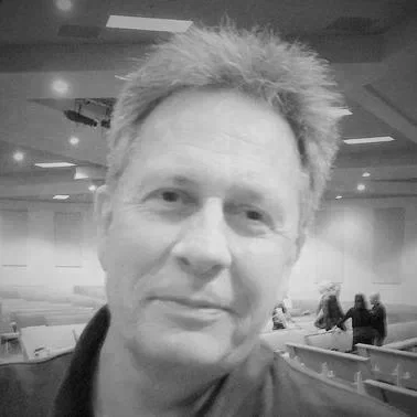 A grayscale photo of a man with short, spiky hair in an indoor setting with people in the background.