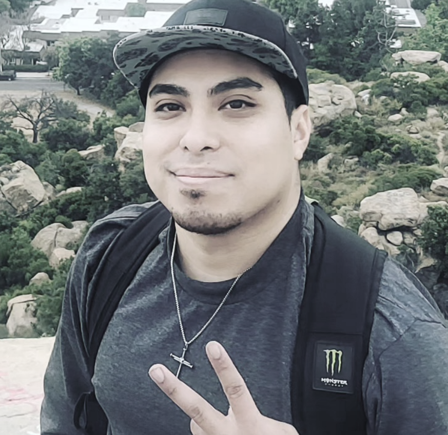 A young man taking a selfie outdoors on a hill or mountain, with rocks and greenery in the background.