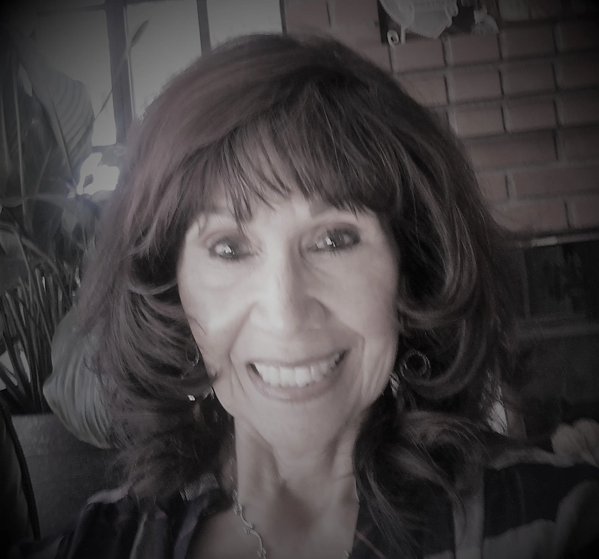 A woman with short, dark hair and light skin smiling widely, showing her teeth, with a background of indoor brick wall and some plants.