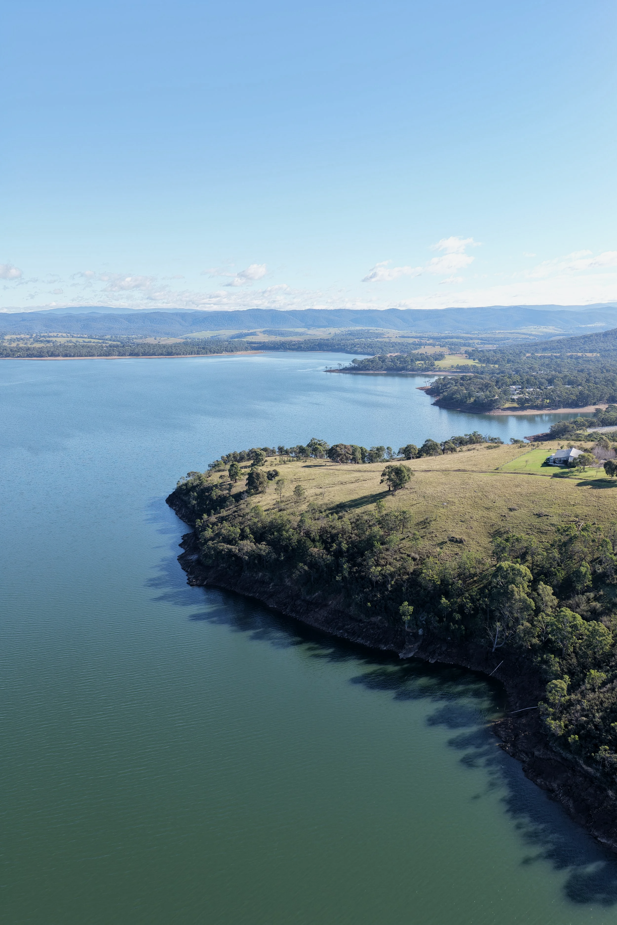 A large body of water, possibly a lake or reservoir, bordered by a green hillside with some scattered trees and buildings, with a backdrop of distant hills or mountains under a partly cloudy sky.