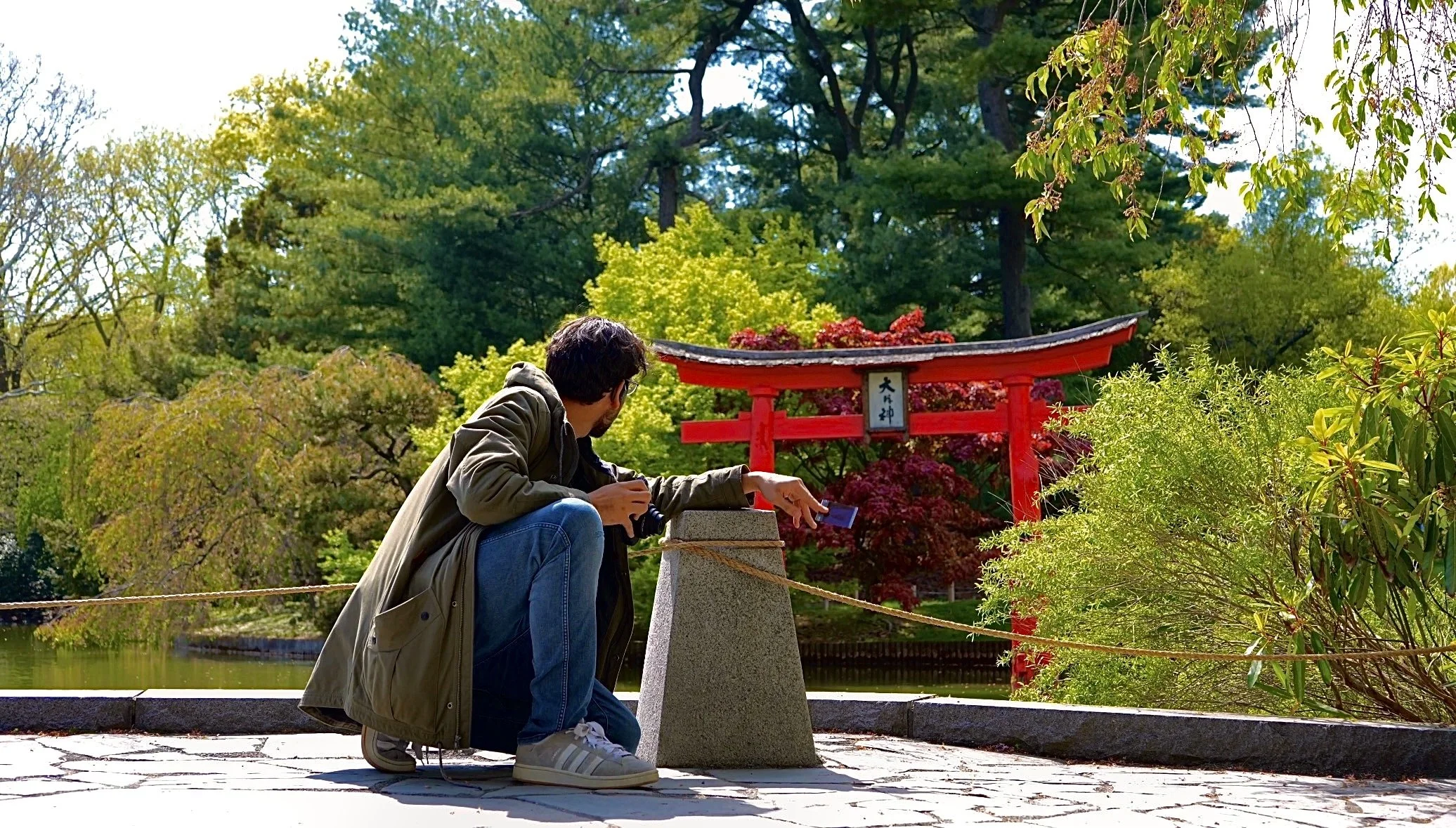 A man kneeling next to a stone lantern in a Japanese garden, with a red torii gate and various green trees and bushes in the background.