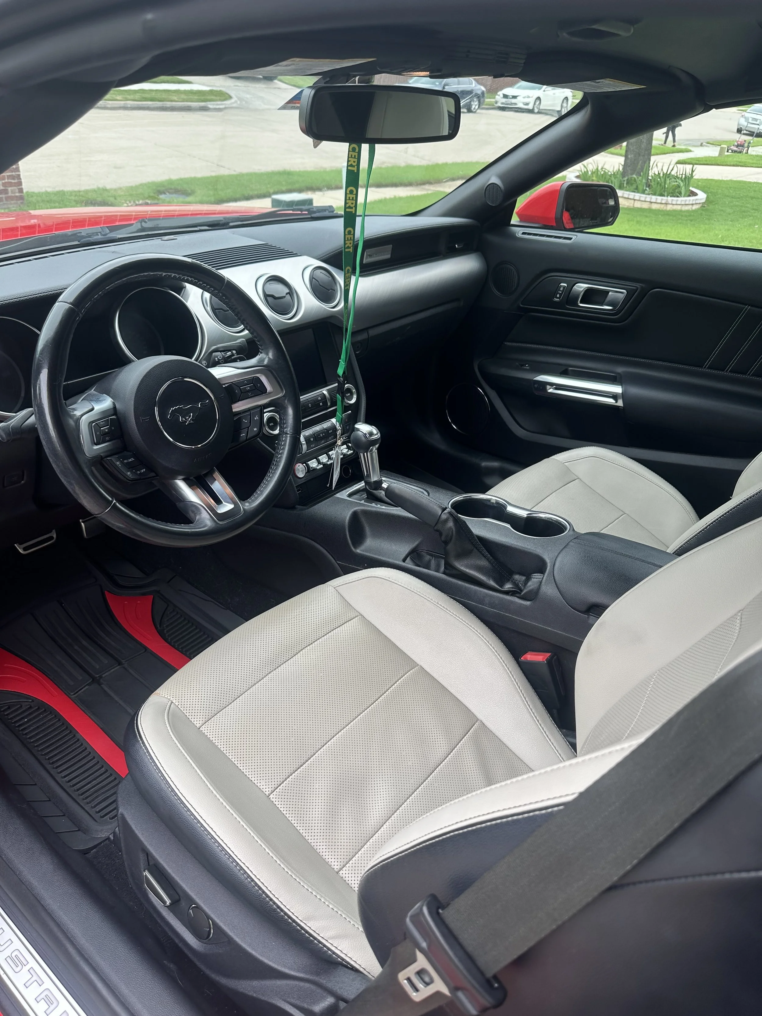 The interior of a black Ford Mustang, showcasing the steering wheel, dashboard, gear shift, beige seats, and the rearview mirror with green and yellow lanyard attached. Outside, a parking lot and some grassy areas are visible.