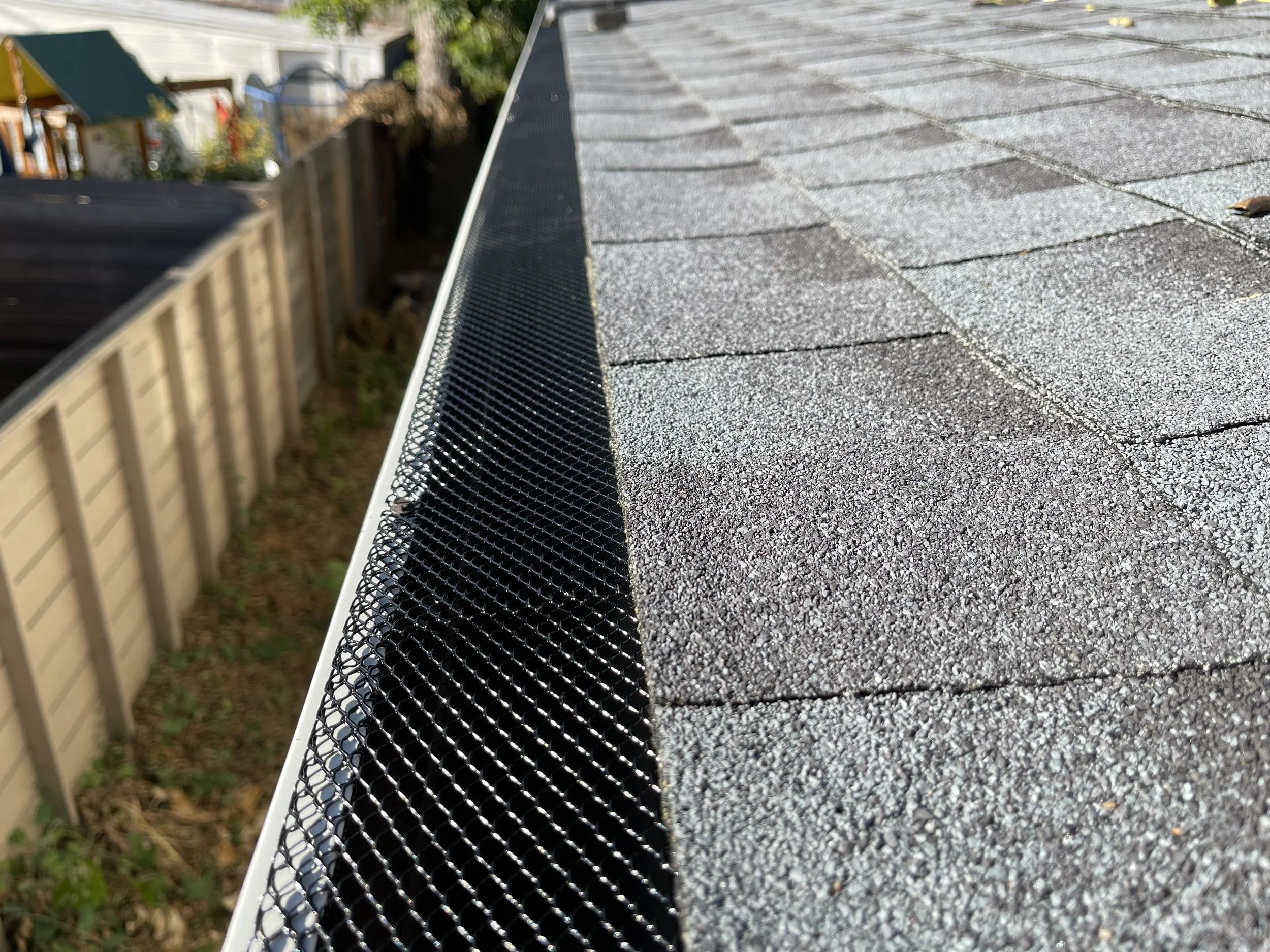Close-up of a black metal rain gutter on a textured gray shingled roof with an out-of-focus background including a wooden fence and trees.