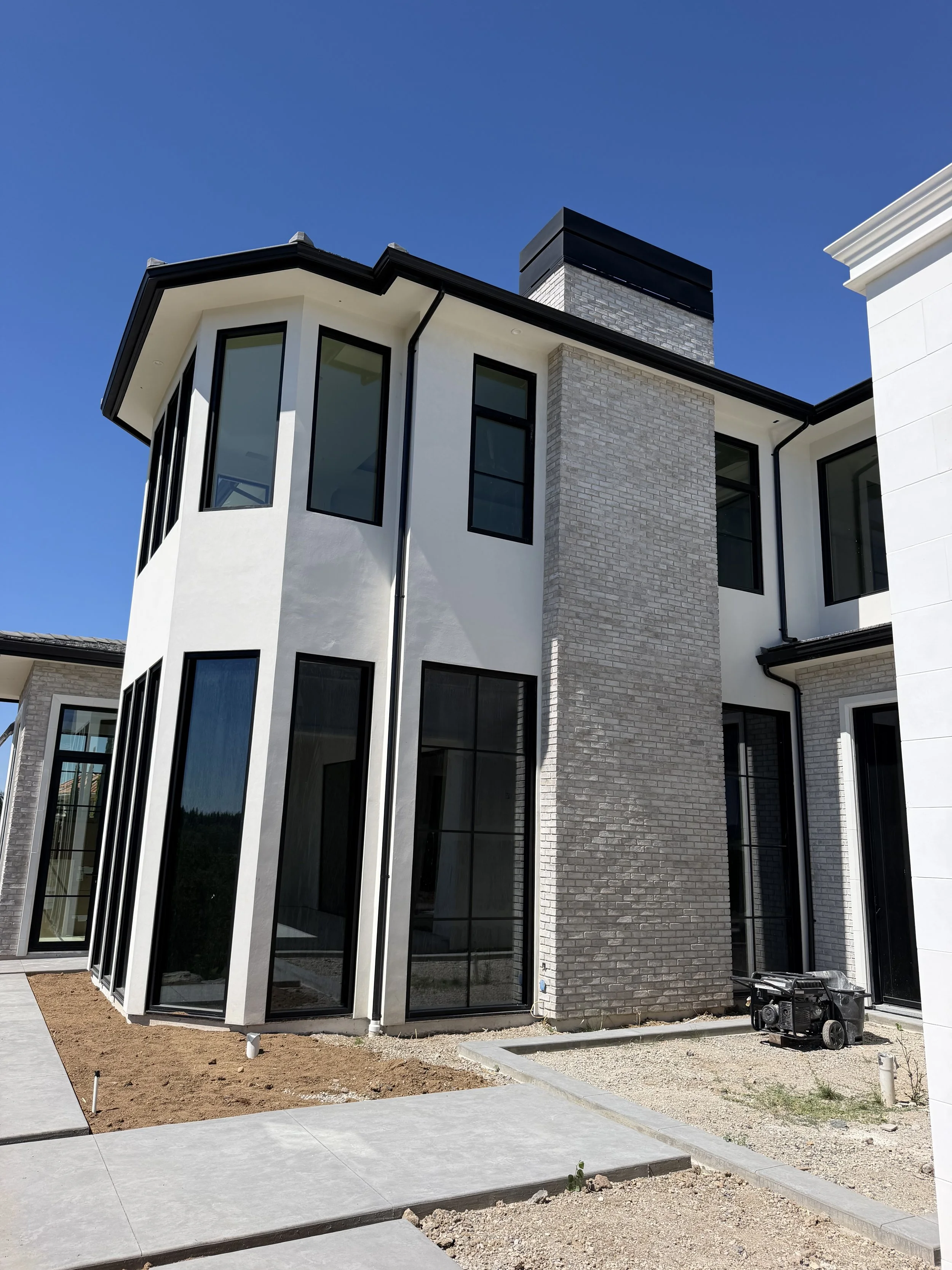 Modern white house with large windows, black trim, a brick chimney, and construction equipment outside during daytime.