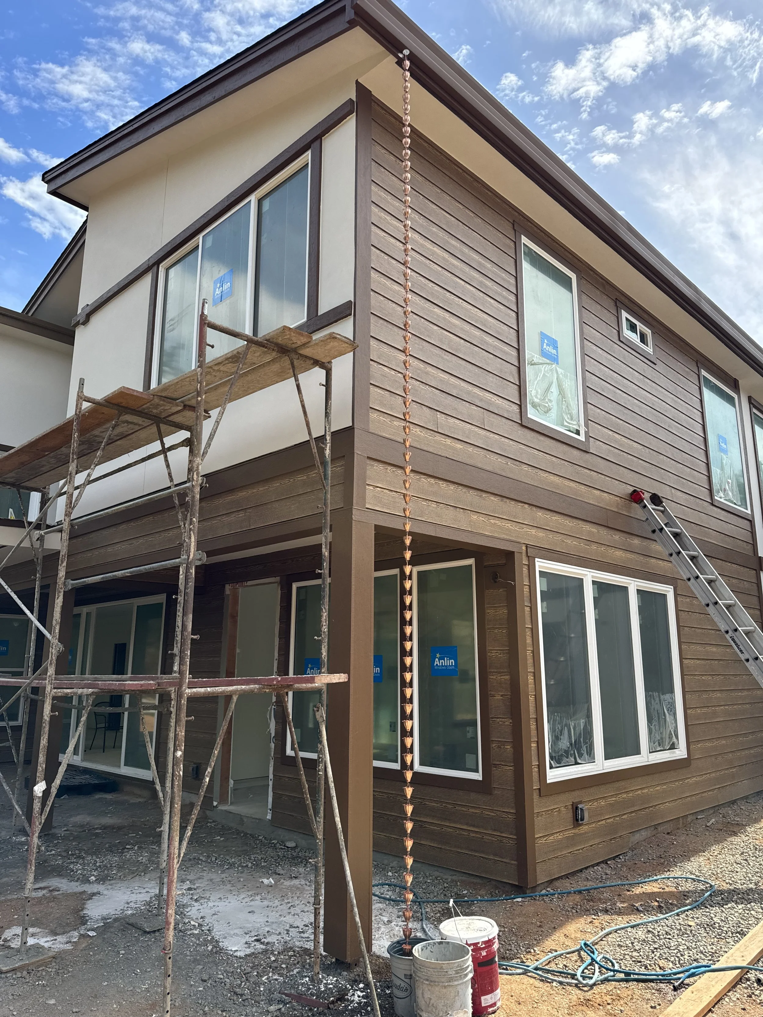 Construction site of a modern two-story house with large windows, wooden exterior siding, a scaffolding and ladder, and construction tools and materials in the foreground.