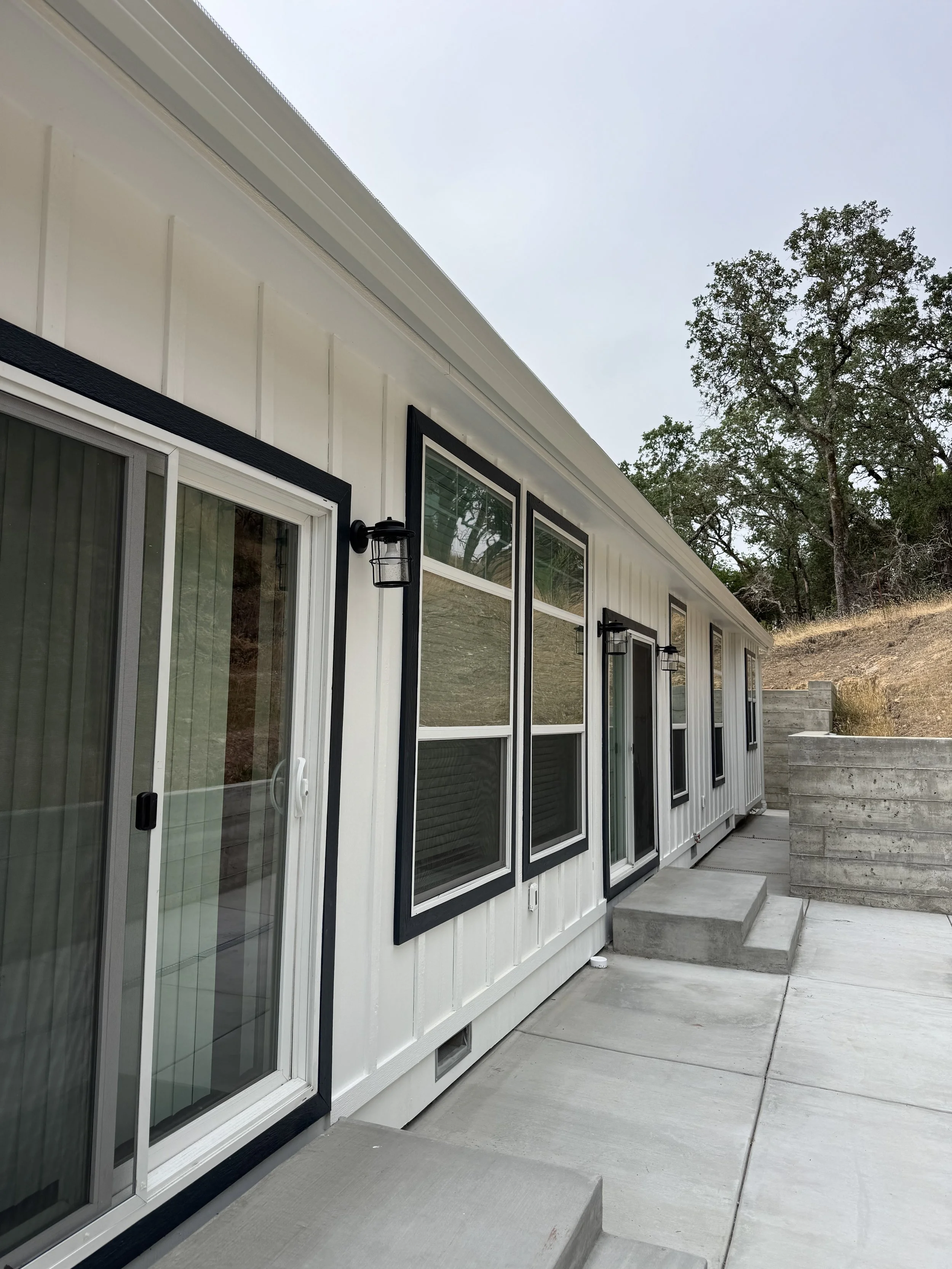 A modern house with several black-framed windows and sliding glass doors, exterior light fixtures, and concrete steps outside. The house has white siding, and there are trees and a grassy hillside in the background.