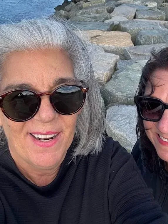 Two women smiling for a selfie by a rocky shoreline, wearing sunglasses.