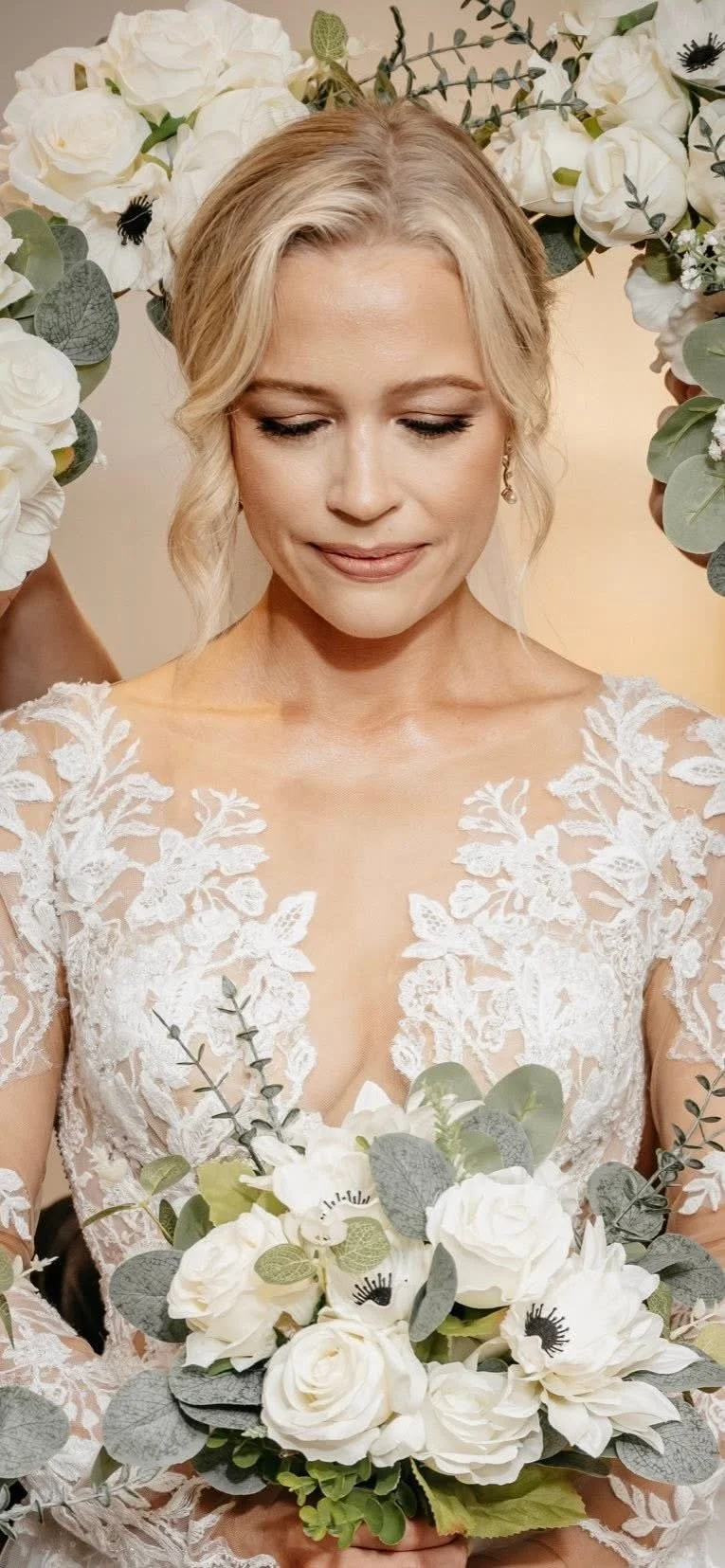 A bride with blonde hair styled in loose waves, wearing a lace wedding dress, holding a bouquet of white roses, anemones, and eucalyptus, surrounded by white flowers and greenery.