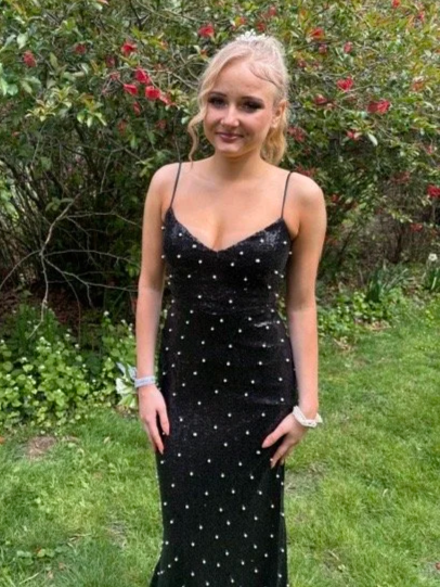 Young woman in a black, spaghetti strap dress with white polka dots, standing outdoors in front of flowering bushes, smiling at the camera.