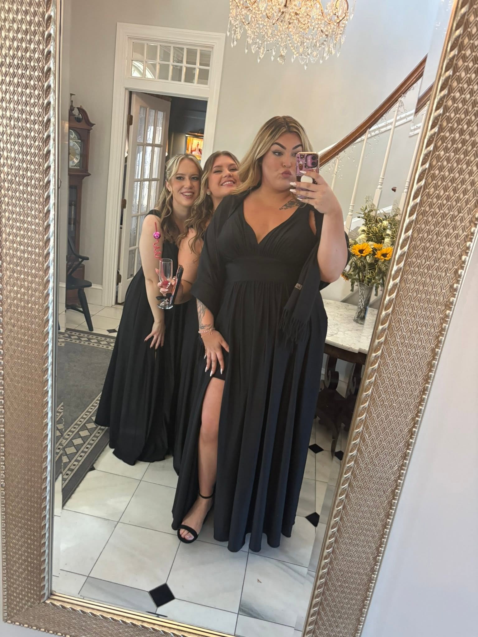 Three women dressed in black gowns taking a mirror selfie at a formal event in a house with white walls, a staircase, and a vase of sunflowers.