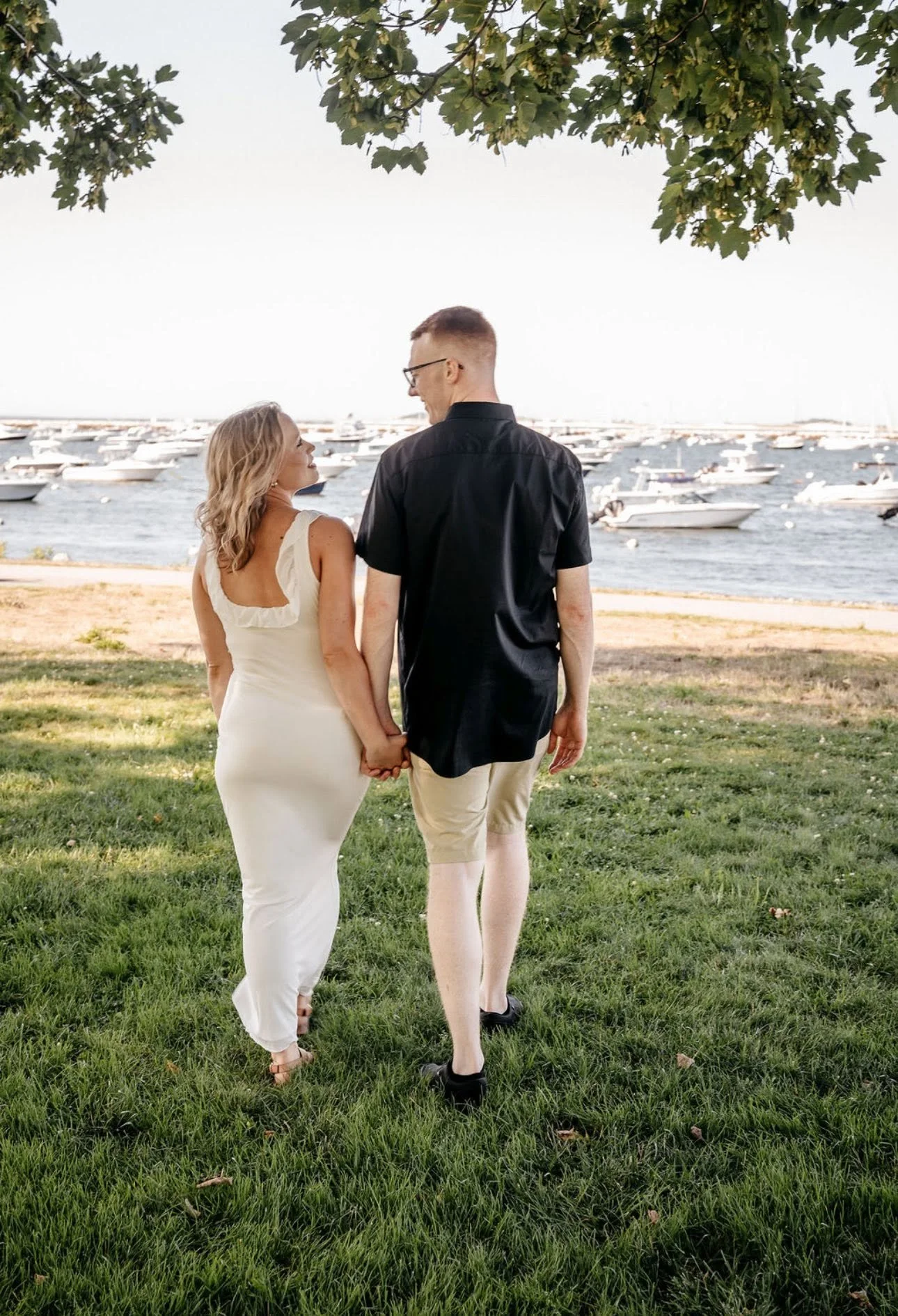A couple holding hands and walking on grass near a marina with boats in the background, under a tree canopy.