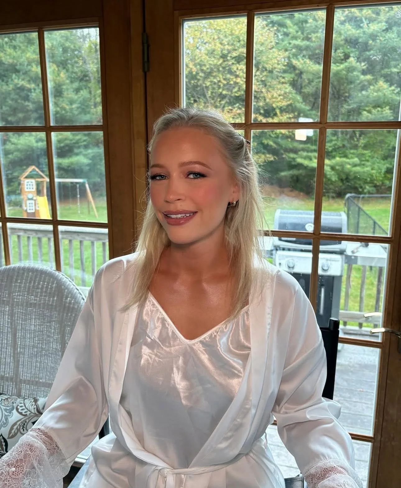 A woman with blonde hair sitting indoors near large windows overlooking a green backyard with trees, a small playhouse, and a grill in the background.