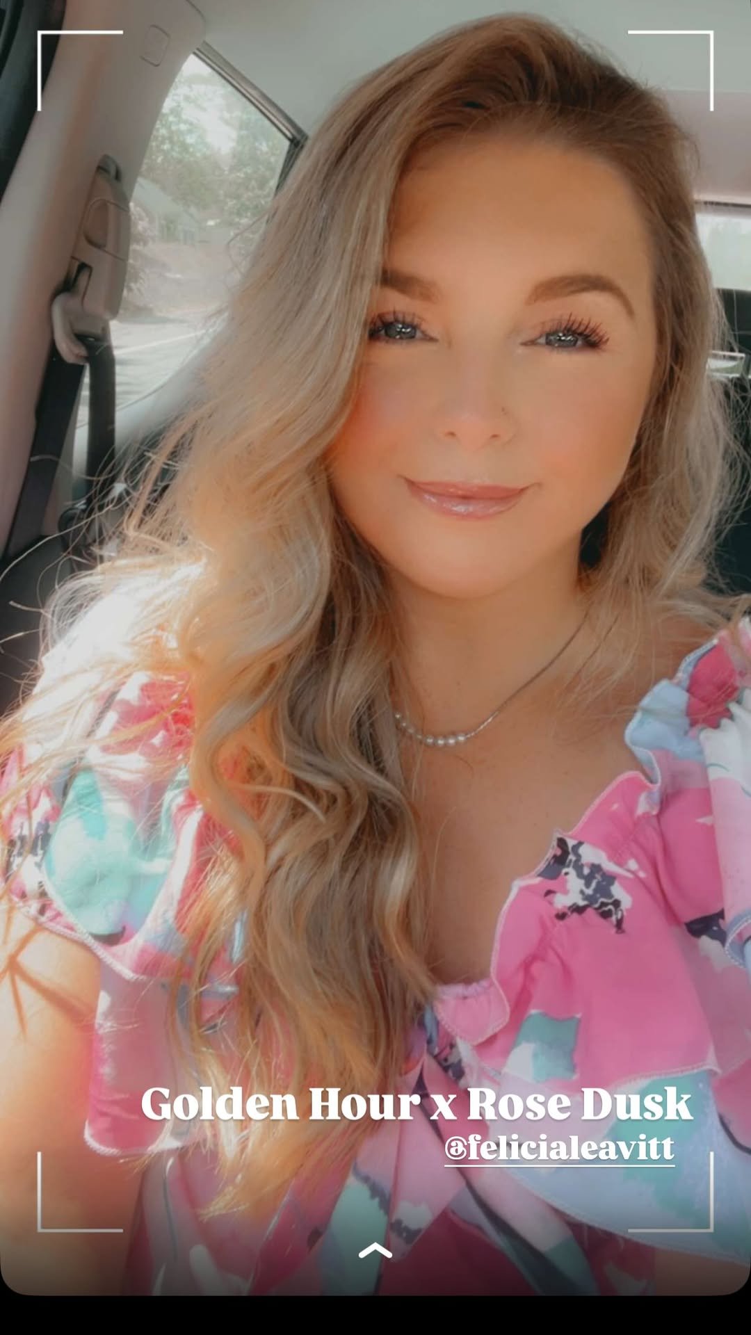 A woman with long, wavy blonde hair sitting in a car, smiling at the camera, wearing a pink floral dress and pearl necklace, with sunlight streaming in.