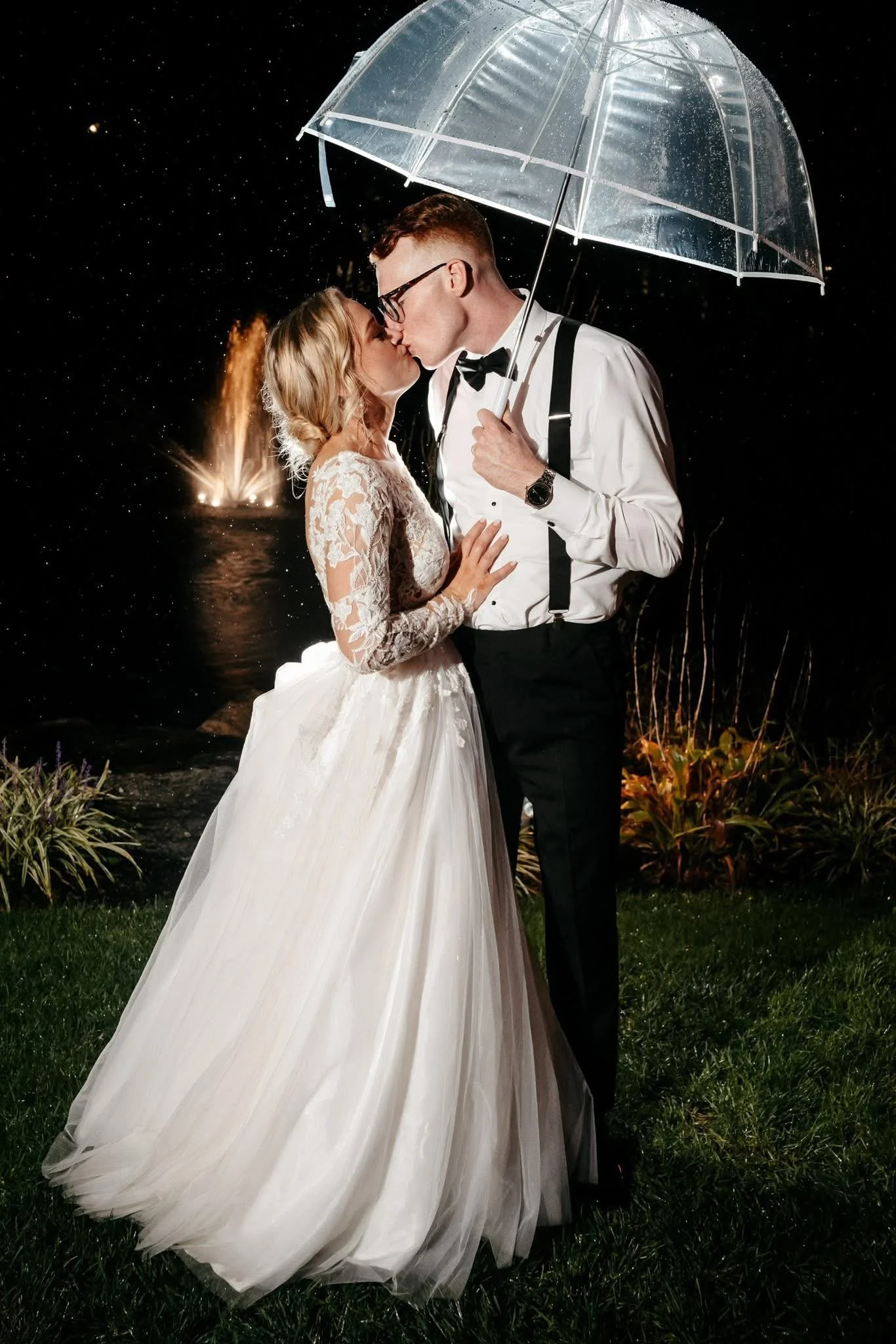 A bride and groom share a kiss under a transparent umbrella during a rainy night. The bride is wearing a white lace wedding dress, and the groom is dressed in a white shirt with black suspenders and bow tie. Fireworks are visible in the background over water, creating a romantic scene.