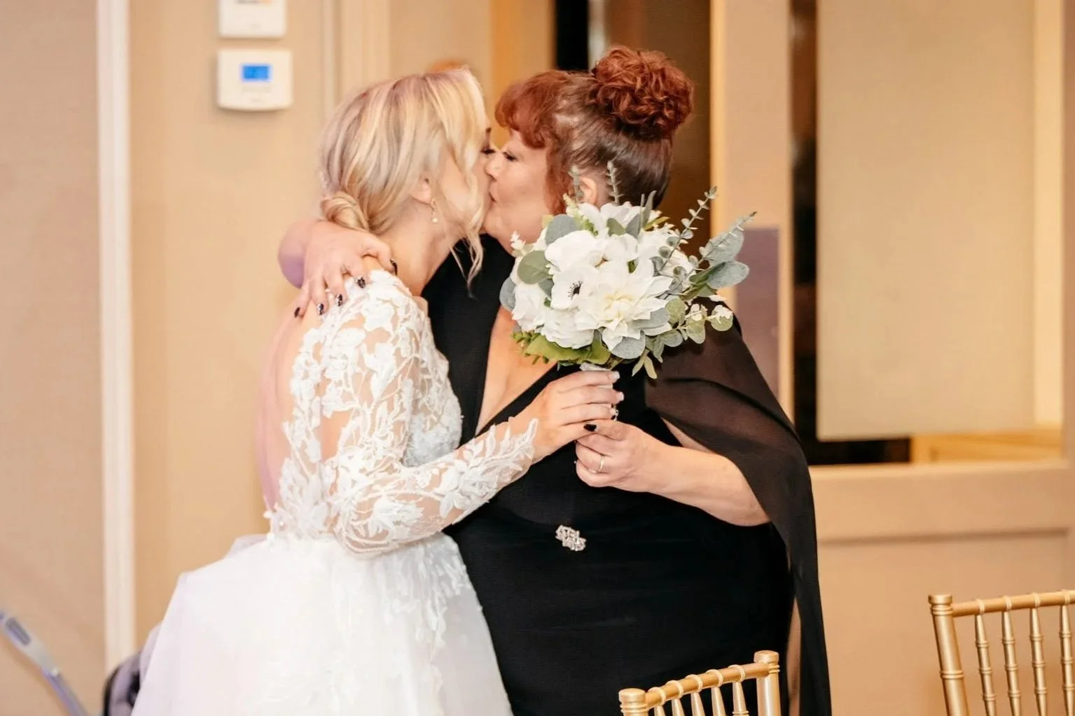 Two women sharing a kiss, one in a wedding dress and the other holding a bouquet of white flowers, at a wedding reception.