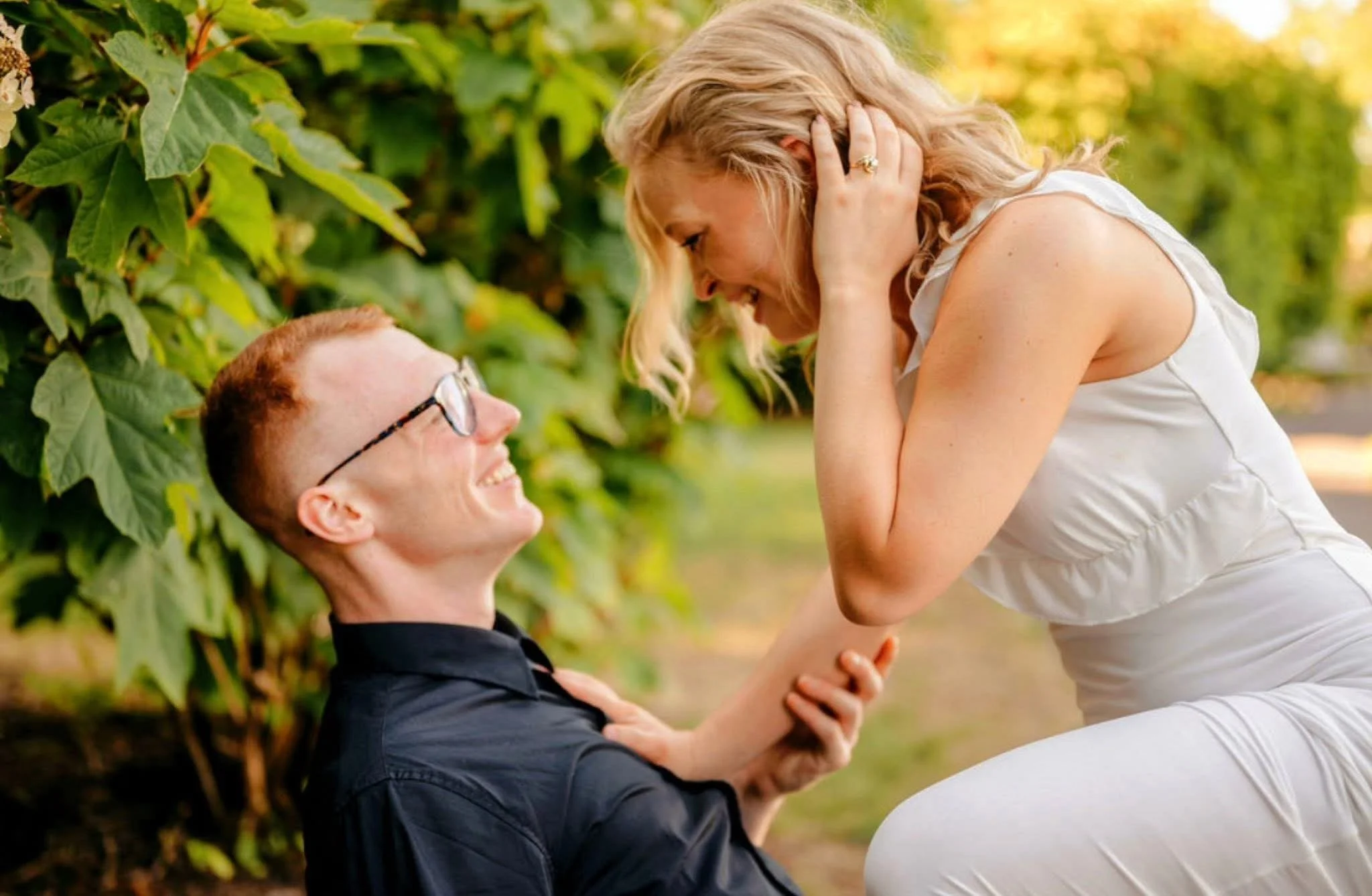 A woman and a man smiling and looking at each other in a garden with green foliage, the woman leaning on the man, holding his face gently, during what appears to be a romantic moment.