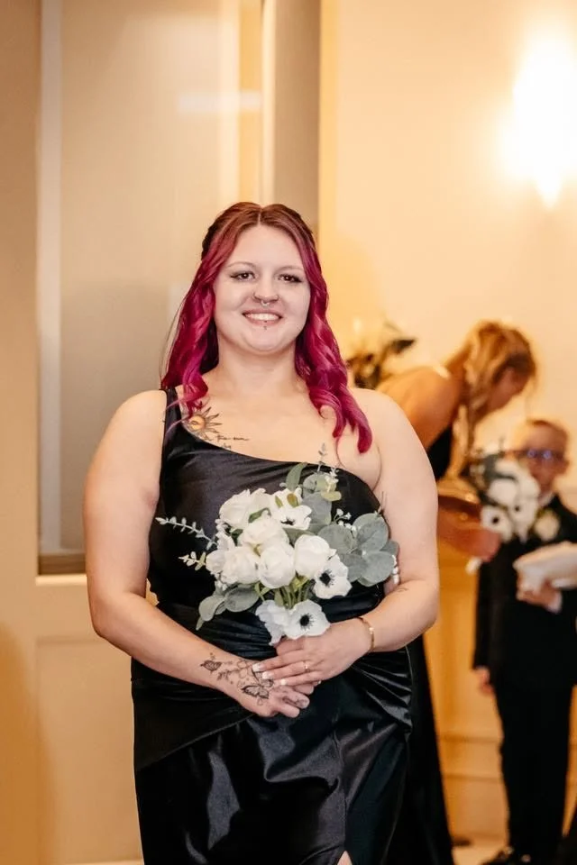 A young woman with pink hair, tattoos, and a nose piercing, holding a bouquet of white flowers, dressed in a black satin gown, standing in a warmly lit interior space.