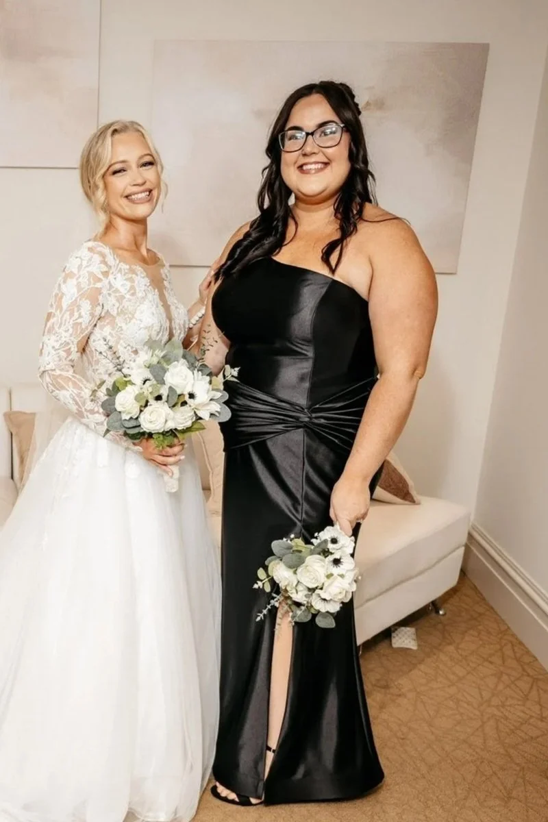 Two women at a wedding, one in a white lace wedding dress holding a bouquet, and the other in a black satin gown holding a bouquet, standing in a room with beige walls and a couch.