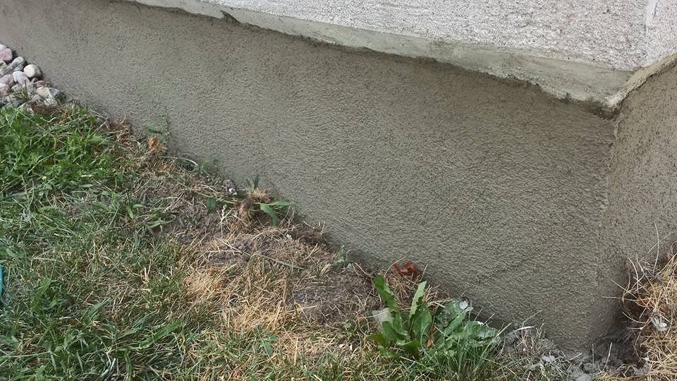 Section of a beige exterior foundation wall with grass and weeds in the foreground.