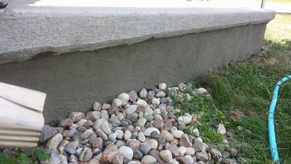 Close-up view of the foundation of a building with a row of small, rounded stones at the base, some green grass, and a blue hose on the right side.