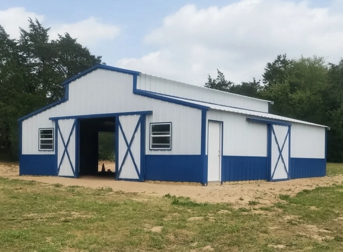 A white and blue barn with double doors on the front, three small windows, and a sloped roof, situated on a patch of grass and dirt with trees and a cloudy sky in the background.
