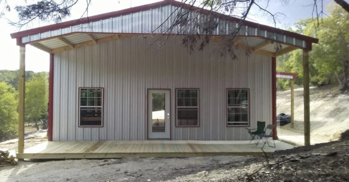 Front view of a metal building with a wooden porch, three windows, a door, two chairs, and an overhang, set on a dirt ground with trees in the background.