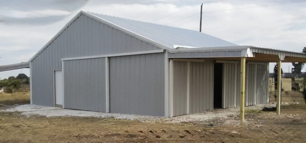 Gray metal storage building on dirt ground with a partially open side and a small covered entryway supported by yellow posts.